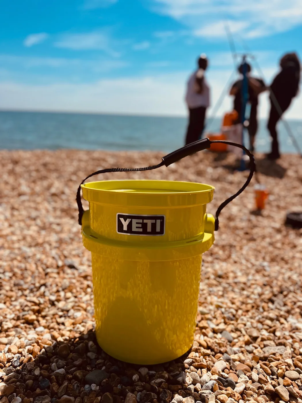 A yellow bucket on a pebble beach with anglers by the sea behind it